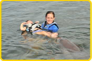 A trainer holds a young guest in a life-vest, while a dolphin noses their feet